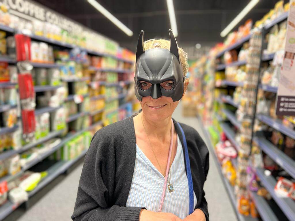 Es zeigt eine Frau in einem Supermarkt. Sie trägt eine Katzenmaske und schmunzelt in die Kamera.